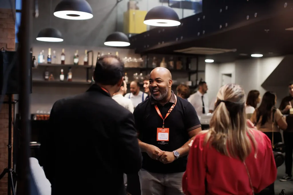 Imagem de um evento social em bar, onde pessoas conversam, destacando um homem sorridente em destaque, criando um ambiente amigável e descontraído.