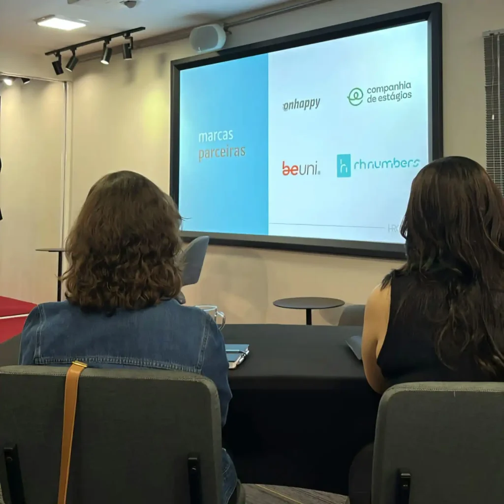 Imagem de uma apresentação com o tema "marcas parceiras" em um evento, mostrando logos de diferentes empresas. Dois participantes assistindo.