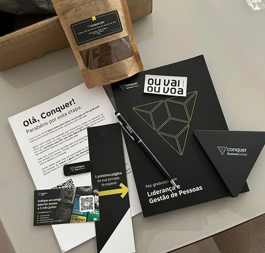 Imagem mostrando materiais da Conquer Business School, incluindo documentos, caneta, embrulho de café e itens promocionais sobre pós-graduação em Liderança e Gestão de Pessoas.