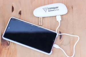 Imagem que mostra um smartphone conectado a um carregador portátil da Conquer Business School, ideal para estudantes e profissionais em movimento.