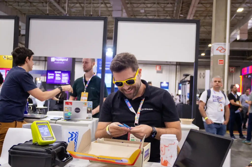 Homem sorridente com óculos escuros interagindo em evento, rodeado por outras pessoas e tecnologia, transmitindo um ambiente de trabalho dinâmico e moderno.
7 ideias de especialistas para destacar seu estande em feiras e conferências